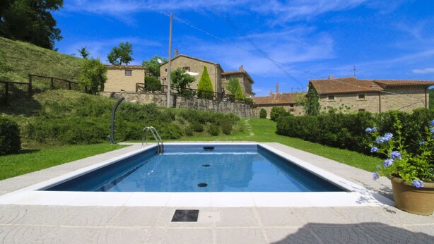 Una piscina con cubierta de piedra, rodeada de follaje y un edificio de piedra al fondo.