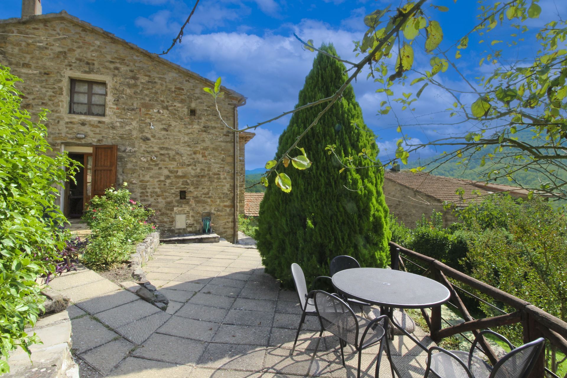 Ein Steingebäude mit Tür und Fenster, ein hoher Baum und ein Tisch mit Stühlen in einem Garten.