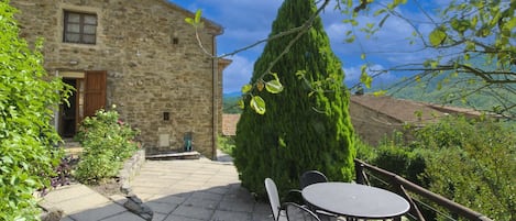 A stone building with a door and window, a tall tree, and a table with chairs in a garden area.