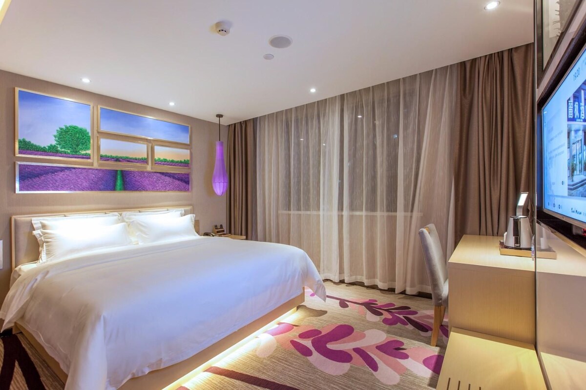 A hotel room with a large bed, a desk with a computer, a TV, and a window with curtains.