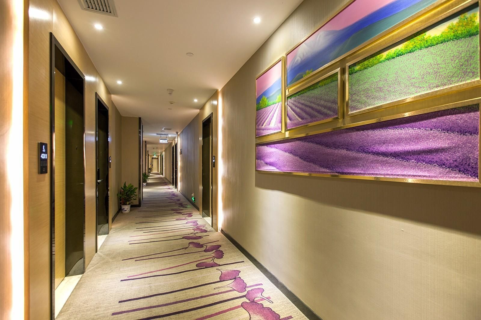 A hotel corridor with a carpeted floor, doors on the left, and wall-mounted artwork on the right.
