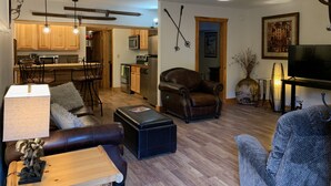 Ein geräumiges Wohnzimmer mit einer Ledersofa, einem Holztisch und einem Fernseher. Im Hintergrund ist eine Küche mit Holzschränken und modernen Geräten zu sehen.