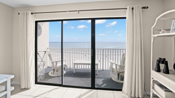 Habitación con balcón que da a una playa, con una mesa y sillas, un estante blanco con binoculares y vistas al mar.