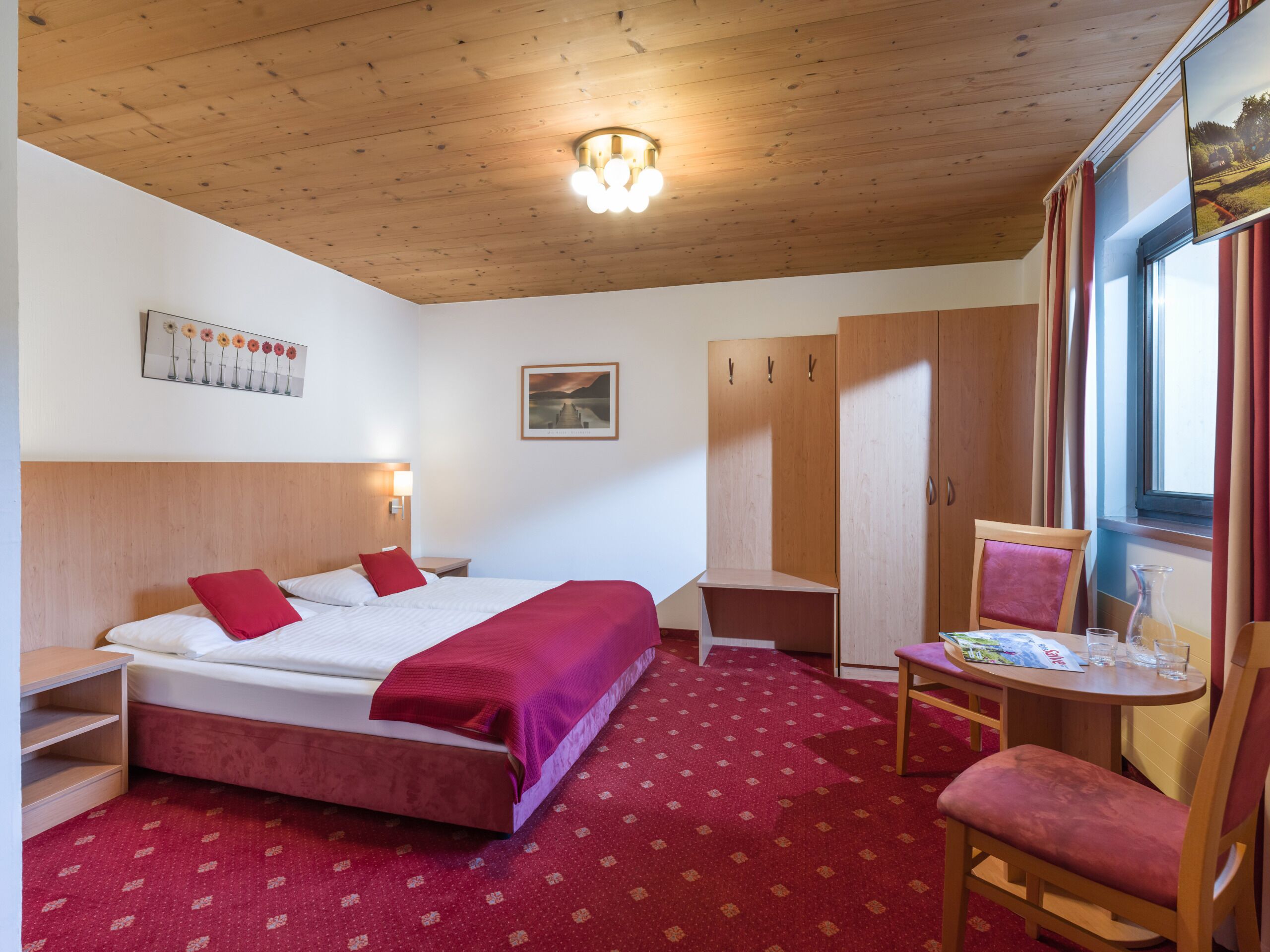 A hotel room with a wooden ceiling, a bed with red bedding, a desk with a chair, and a window with curtains.