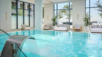Piscina interior con vistas despejadas al mar a través de amplios ventanales, rodeada de zonas de descanso y plantas en macetas.