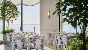 A dining area with tables set for four, chairs with a unique pattern, and large windows overlooking a body of water. - The Pearle Hotel & Spa, Autograph Collection (Burlington)