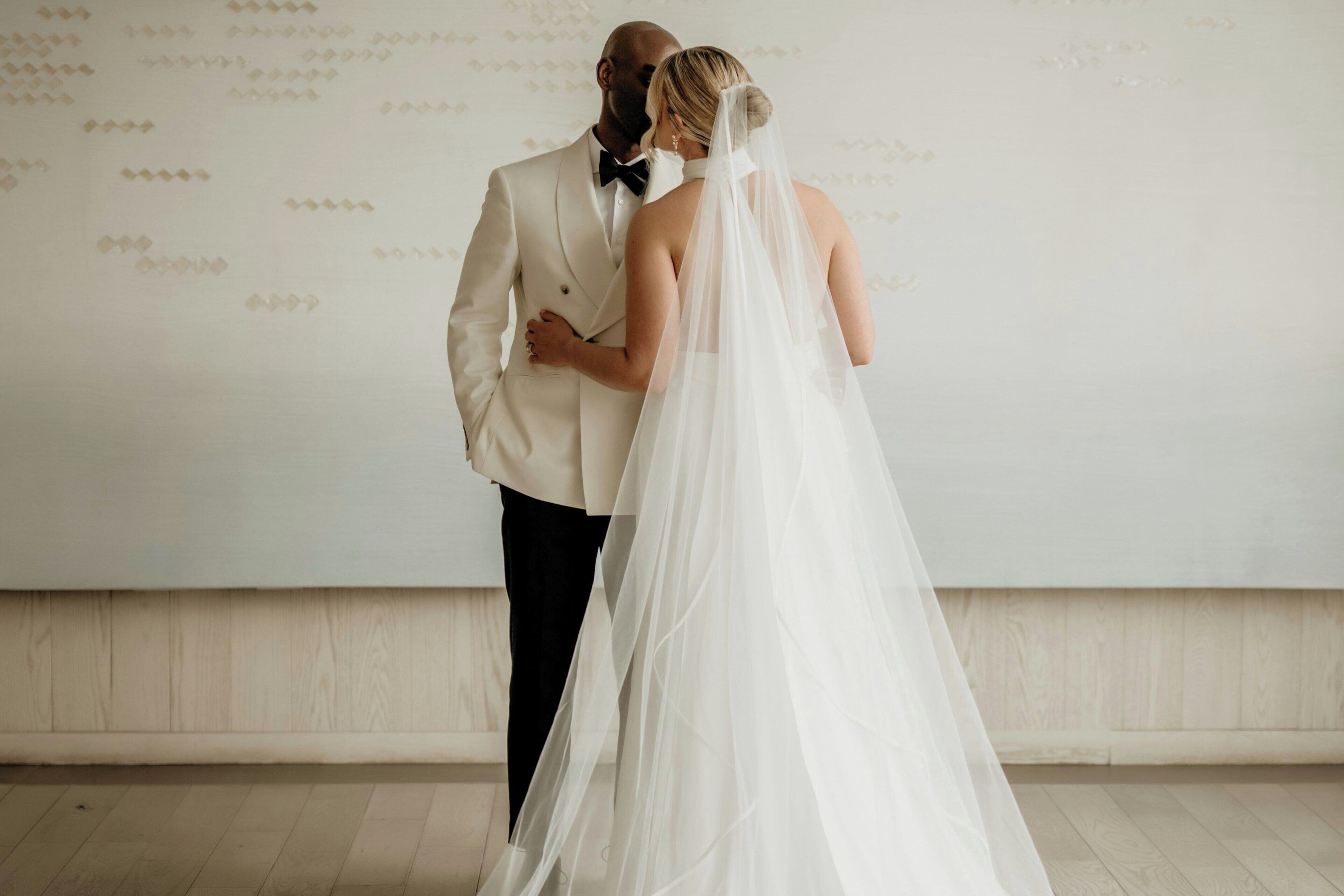 A couple in wedding attire standing in a room with a plain wall and wooden flooring.