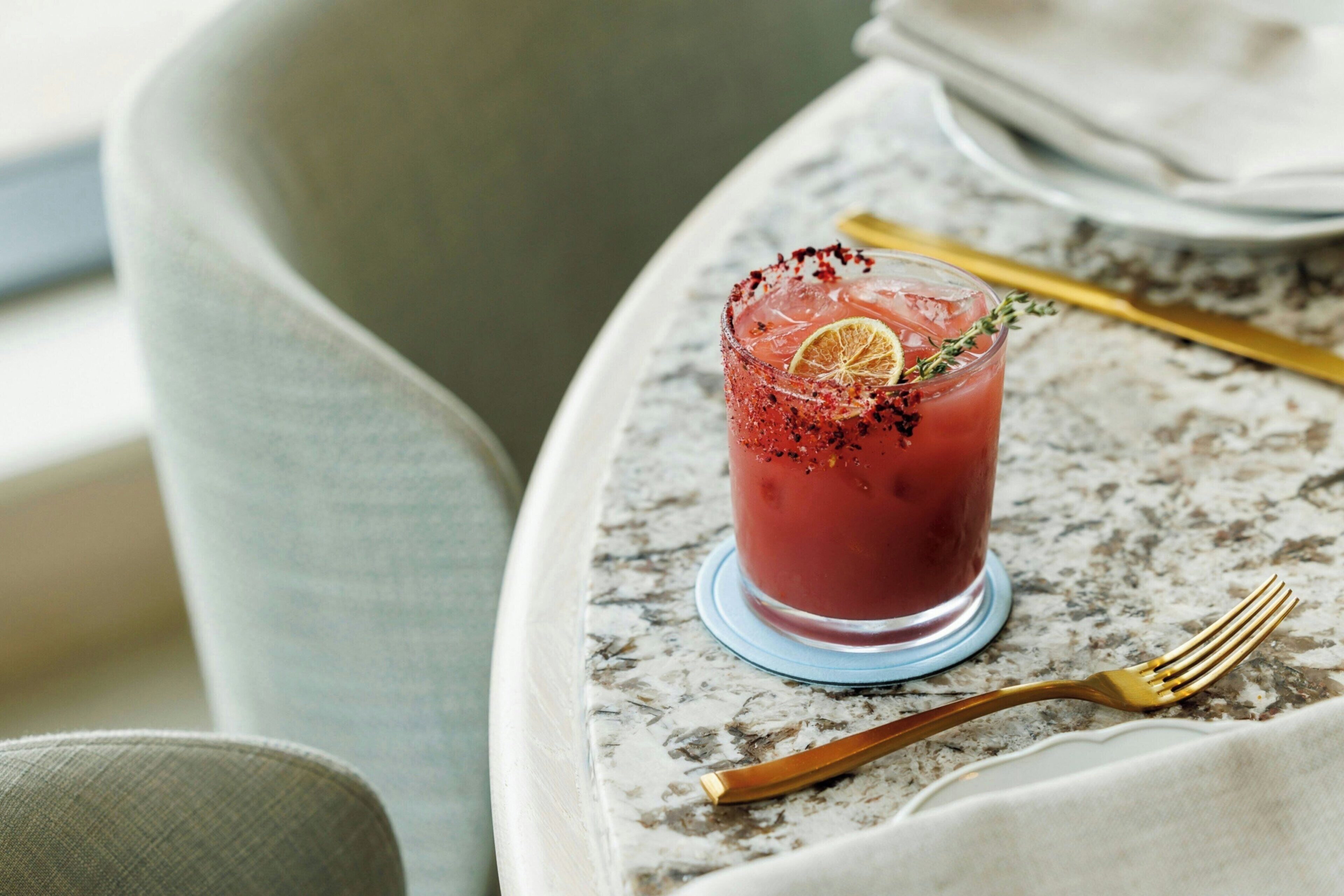 A glass of red drink with a lemon slice and a sprig of thyme on a marble table.