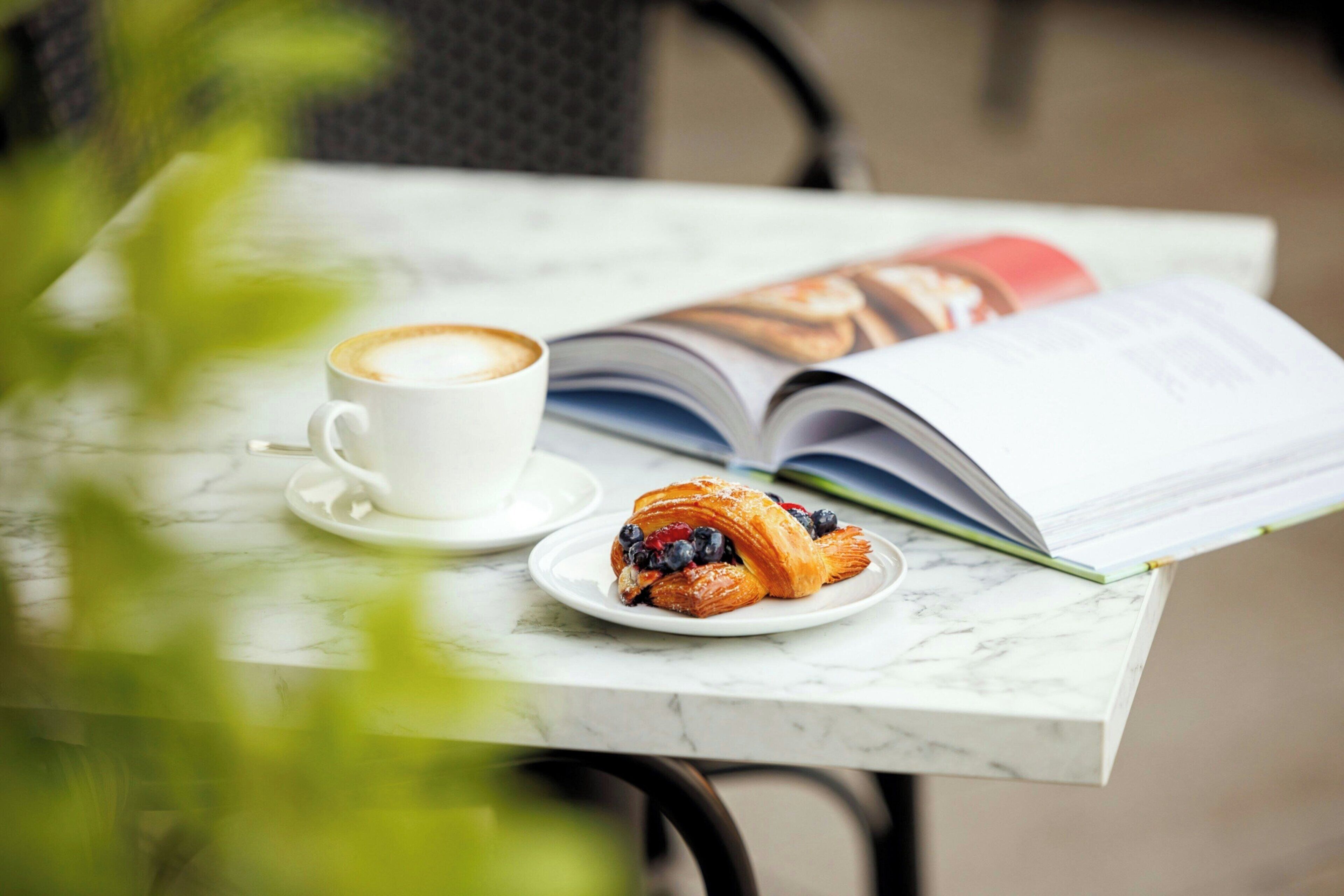 Una mesa con una taza de café, un plato con un pastel y una revista abierta.