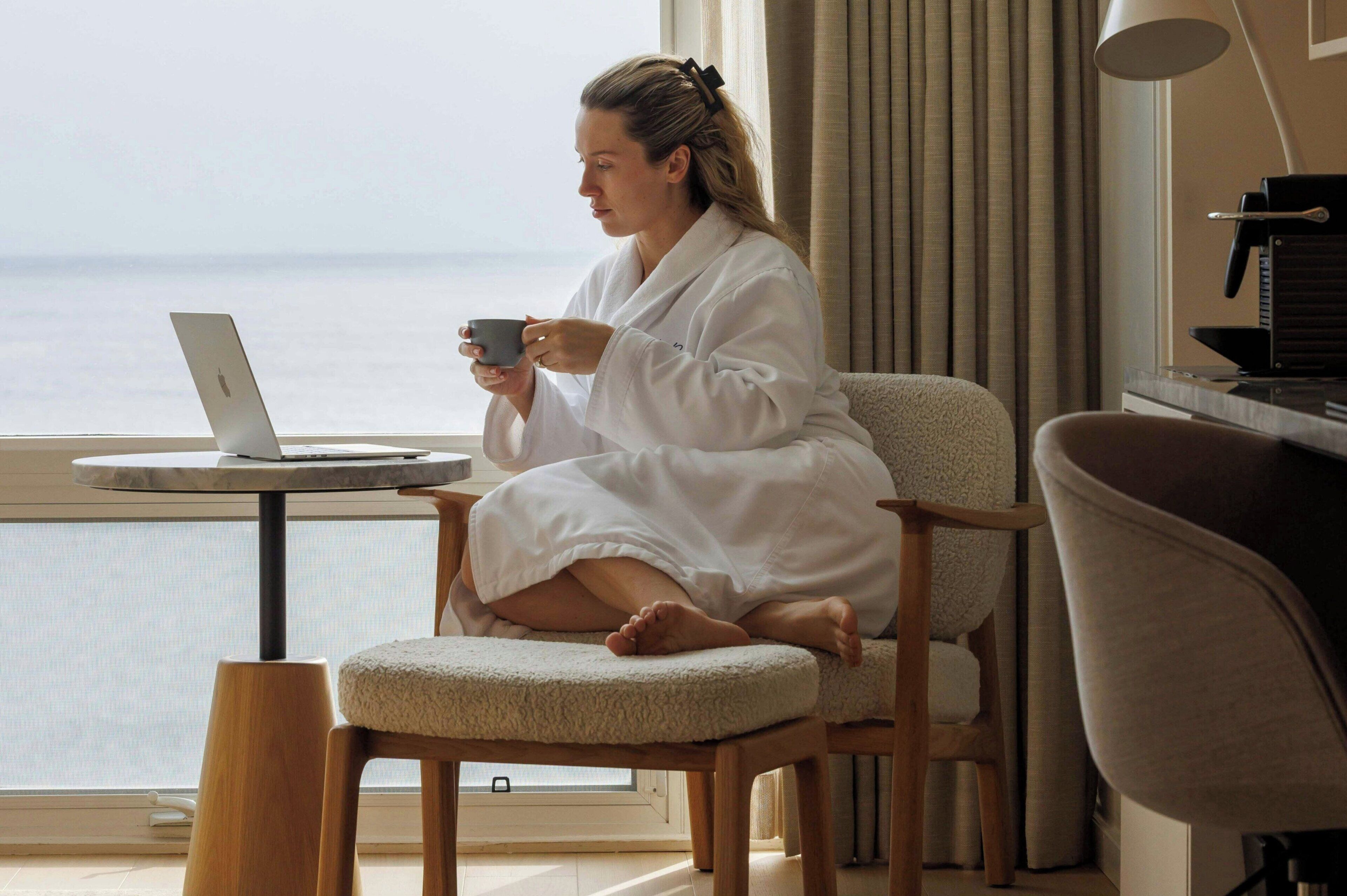 Une personne vêtue d’un peignoir blanc est assise sur une chaise près d’une fenêtre, un ordinateur portable et une tasse sont posés à côté d’elle.