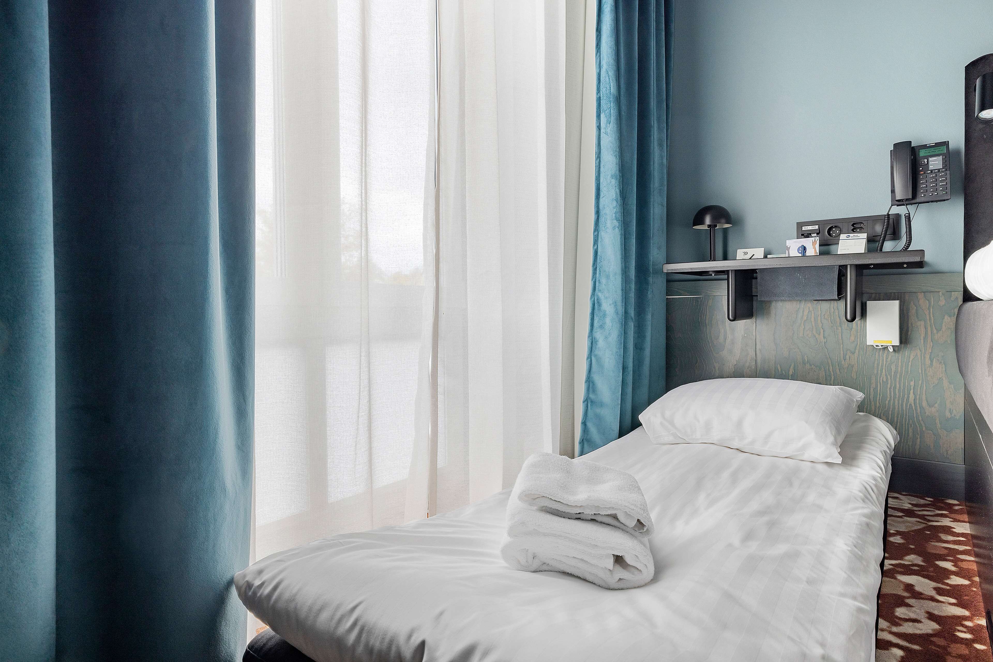 A hotel room with a bed, a desk with a telephone, and a window with curtains.