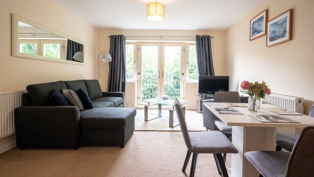 A modern living room with a sofa, a glass-top coffee table, a dining table with chairs, a TV, and a vase with flowers. - Apartment 4 (Worksop)