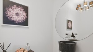Un baño con un espejo grande, un lavamanos, un florero con flores y un cuadro en la pared.