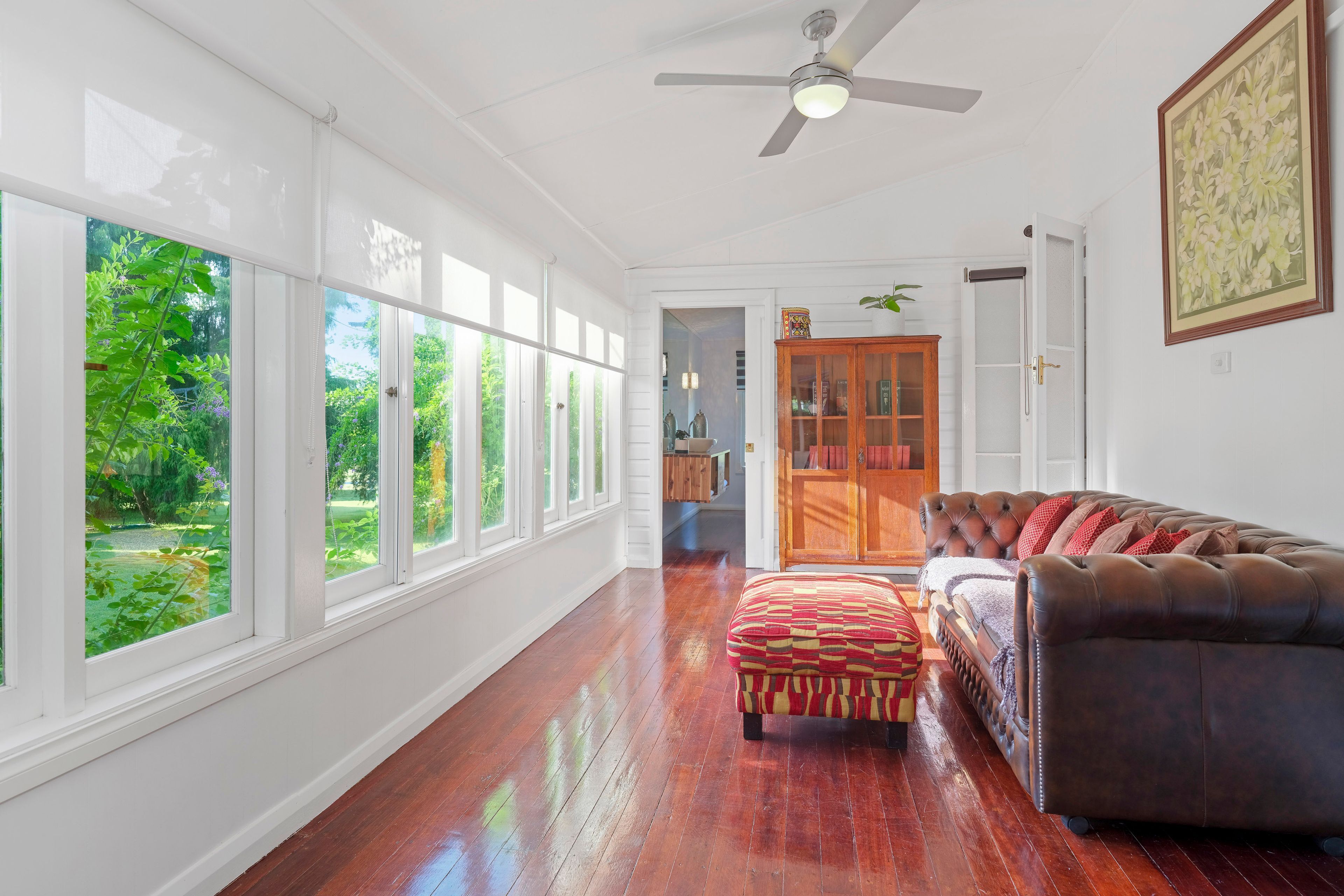 A spacious room with a large window, a ceiling fan, a leather sofa, and a wooden cabinet.