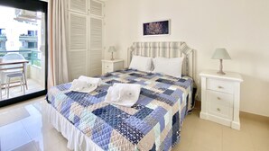 A bedroom with a bed, two bedside tables, a window with curtains, and a view of buildings outside. - Tália XI - Near the beach - Vilamoura (Vilamoura)
