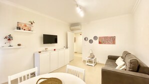 A living room with a white dining table, a grey sofa, a TV mounted on the wall, and a small white cabinet. - Tália XI - Near the beach - Vilamoura (Vilamoura)