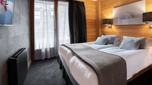 Una habitación de hotel moderna con una cama grande, cabecera de madera, un ventanal con cortinas, un radiador y un televisor montado en la pared.