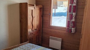 A small, cozy room with a wooden wardrobe, a bed with a patterned bedspread, a radiator, and a window with a view of a snowy landscape. - Chalet in Pra Loup 1500 with 4 bedrooms for 12 people (Pra-Loup)