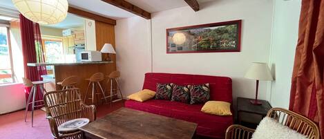 Una sala de estar acogedora con un sofá rojo, una mesa de centro de madera, sillas de mimbre y un tapiz estampado. Se ve una cocina al fondo, con un microondas y taburetes de bar.