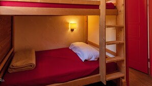 Una cama litera con colchón rojo, almohada blanca y escalera de madera. Encima de la cama, una lámpara fijada a la pared.