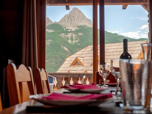 A dining table with plates, glasses, and a bottle, set against a mountain view through a window.