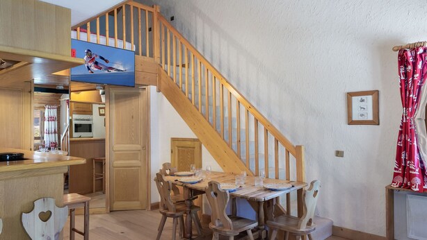 Une cuisine chaleureuse, meublée de meubles en bois, avec une table à manger pour quatre personnes et un escalier en bois menant à l’étage supérieur.