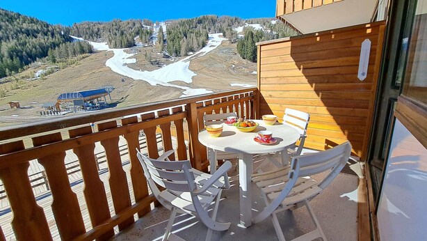 A balcony with a table and chairs, overlooking a snowy mountain landscape with a ski lift and buildings. - Renovated studio at the foot of the slopes in Pra-Loup, ideal for 4 people (Pra-Loup)