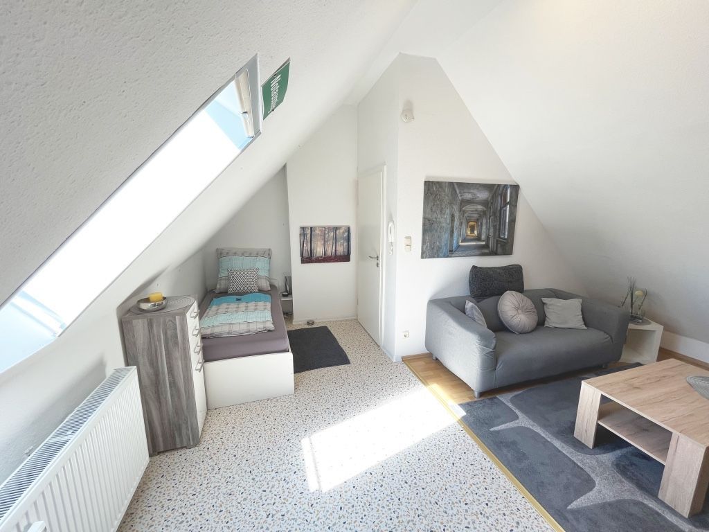 Ein Loft-Zimmer mit schrägem Dach, einem grauen Sofa, einem Holztisch und einem Bild an der Wand.
