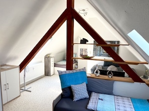 A loft bedroom with a sloped ceiling, a bed with blue and patterned pillows, a wooden shelf with decorative items, and a radiator.
