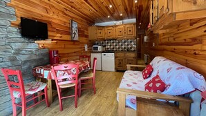 A cozy cabin interior with a stone wall, wooden ceiling, a dining area with a table and chairs, a sofa, and a kitchen with wooden cabinets and appliances.