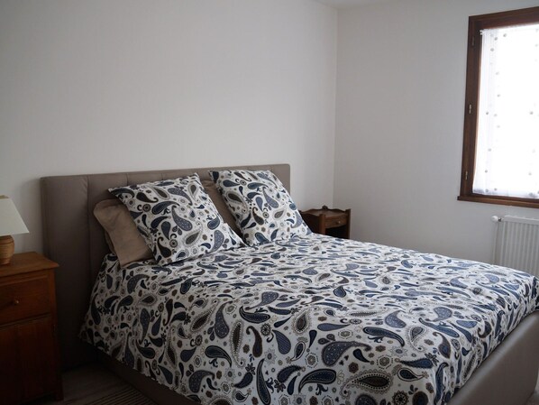 A bedroom with a bed, a nightstand, a lamp, and a window with blinds. - Renovated T3 Apartment in Barcelonnette Center, sleeps 6, balcony (Barcelonnette)