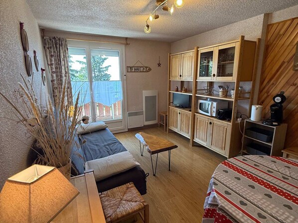 A compact room with a bed, a small table, a kitchenette, and a window with curtains.