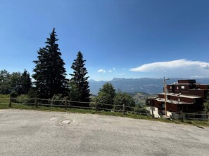 A paved area with a wooden fence, a building with a flat roof, and a mountain range in the background.