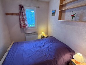 A bedroom with a bed, a nightstand with a lamp, a window with curtains, a radiator, and a small shelf with a flower.
