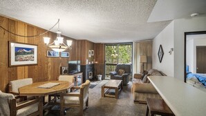 A cozy living room with a wooden wall, a fireplace, a television, and a large window. - Crestview 29:  Walk to the Village, Near Blue Line (Mammoth Lakes)