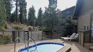 A hot tub on a wooden deck with a metal railing, surrounded by trees and a building with a balcony. - Crestview 29:  Walk to the Village, Near Blue Line (Mammoth Lakes)