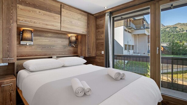 A modern hotel room with a wooden headboard, a large bed with white linens, two wall-mounted lamps, and a balcony with a mountain view.