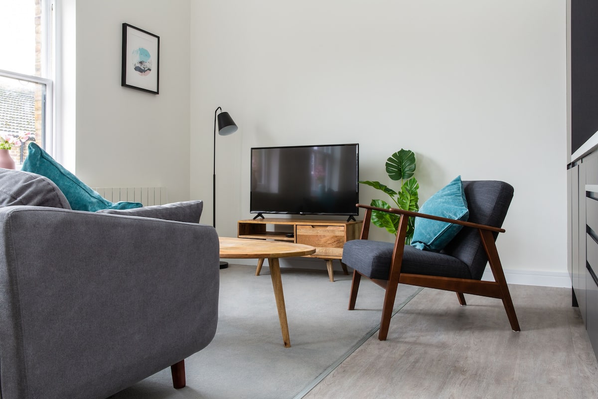 Ein modernes Wohnzimmer mit grauem Sofa, einem Holztisch, einem dunkelgrauen Sessel, einem Flachbildfernseher auf einem Holzständer und einer großen Zimmerpflanze.