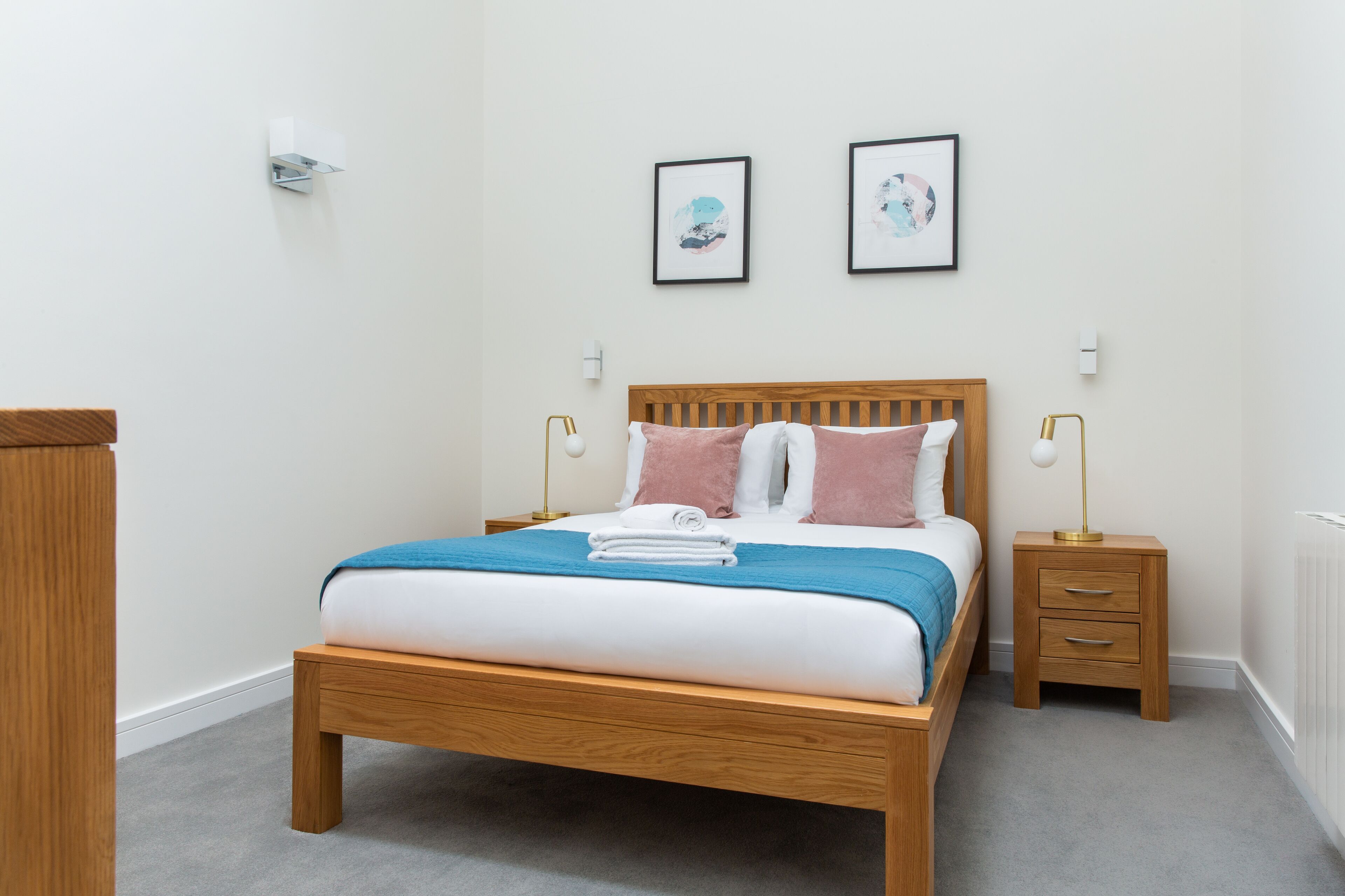 Un lit bien fait, avec une couverture bleue, une tête de lit en bois et deux tableaux encadrés accrochés au mur.
