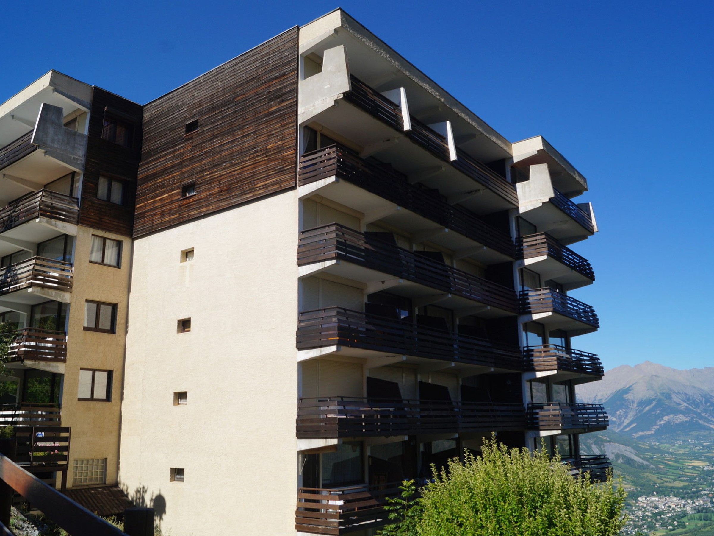 A multi-story residential building with balconies, a flat roof, and a view of mountains in the background.