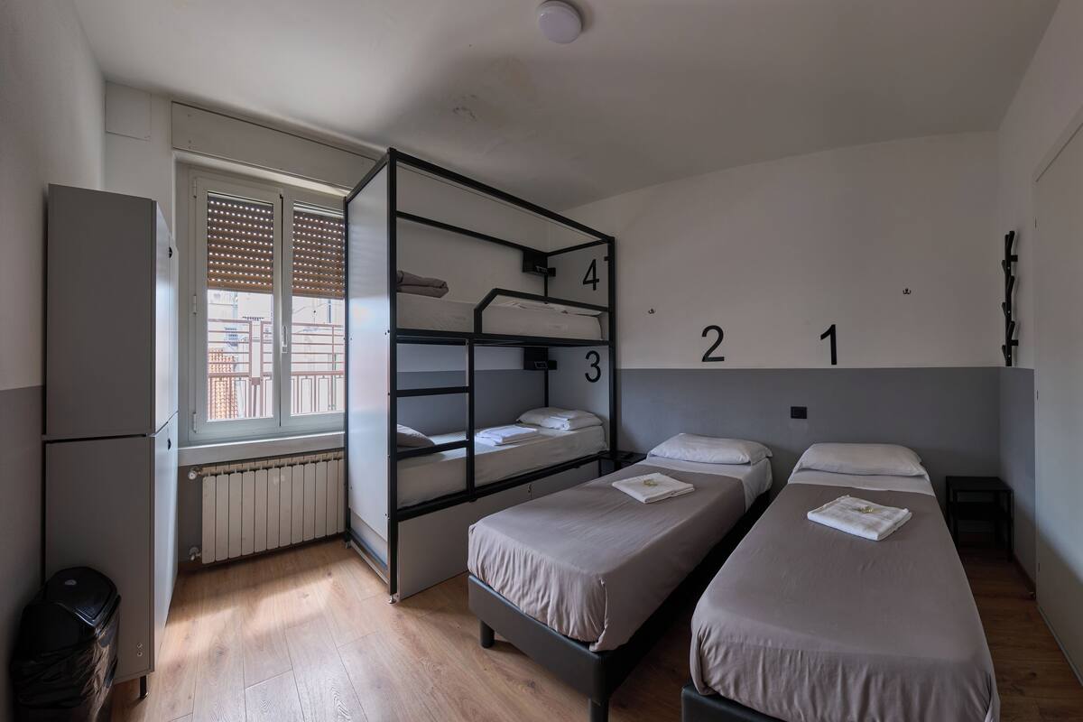 A dormitory room with bunk beds, numbered beds, a radiator, a window with blinds, and a refrigerator.