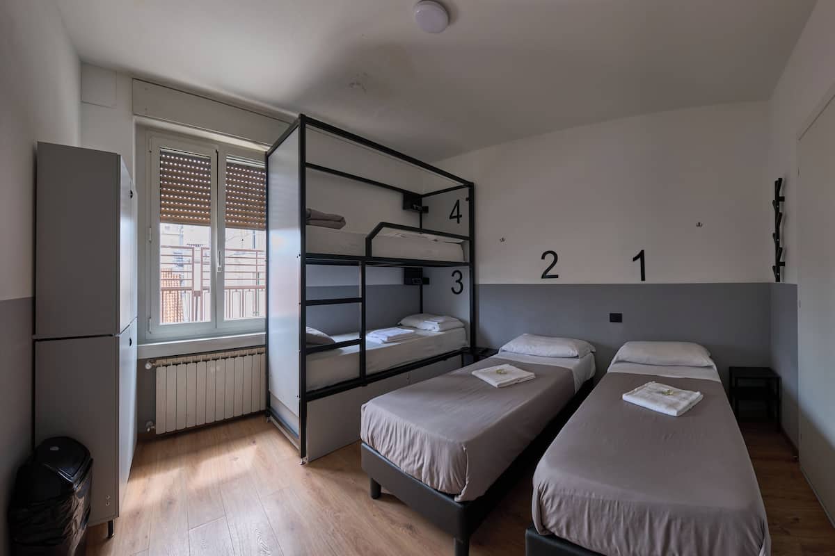 A dormitory room with bunk beds, numbered beds, a radiator, a window with blinds, and a refrigerator.
