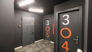 Un couloir avec les portes numérotées 304 et 305, un panneau indiquant les toilettes et un panneau rouge signalant la sortie de secours.