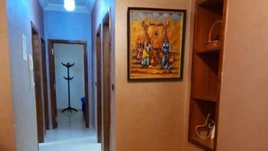 A hallway with a tiled floor, a coat rack, a painting on the wall, and a small wooden stool. - Residence Annaser 3 Agadir (Hay Essalam,Agadir)