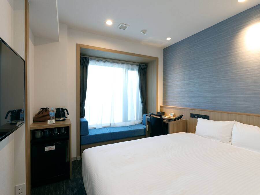 Hotelkamer met een groot bed, een bureau met stoel, een kleine kitchenette en een raam met gordijnen.