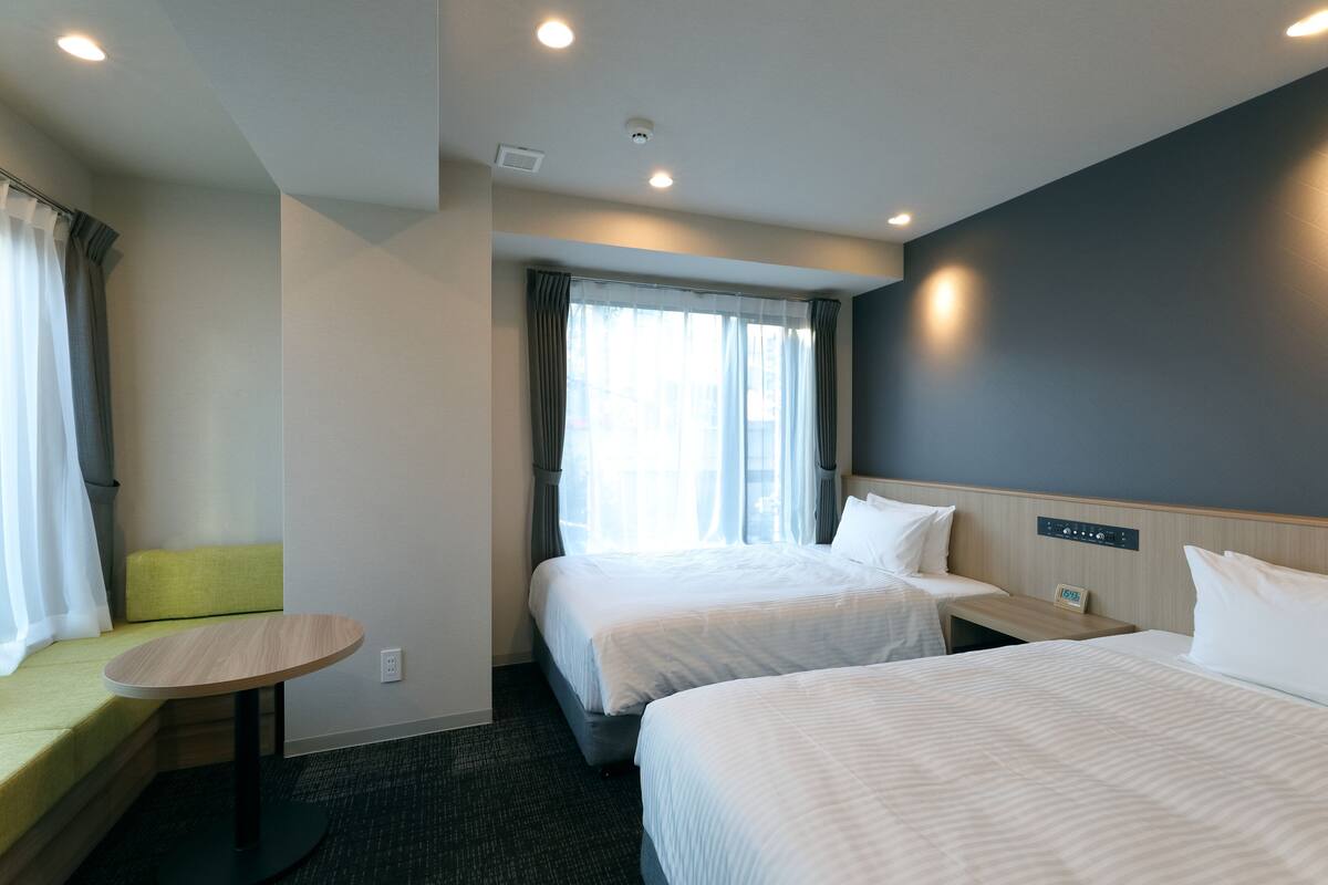 A hotel room with two beds, a small table, a bench, and a window with curtains.