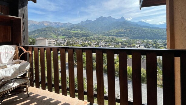 A balcony with a wooden railing overlooking a mountainous landscape with a town below. - Mini T2 Duplex in Barcelonnette, perfect for 4 (Barcelonnette)