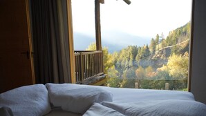A bed with white bedding, a wooden headboard, and a view of a forested hillside through a window. - Holiday Home "Bergsuite" with Mountain View, Balcony, Garden & Wi-Fi (St.Martin in Passeier)