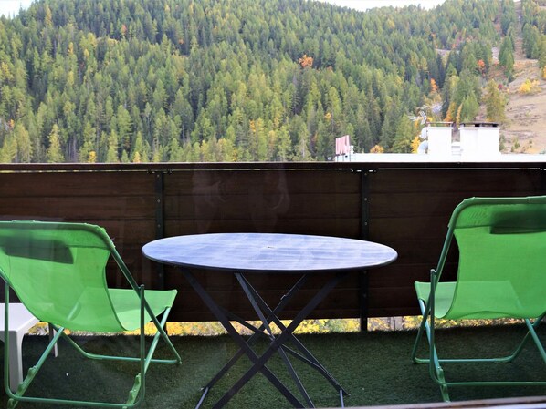 A balcony with a round table and two green chairs, overlooking a forested hillside.