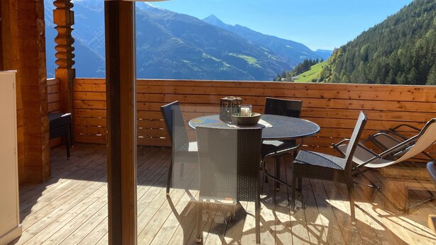 Une terrasse en bois avec une table et des chaises, et une porte vitrée donnant sur une vue sur les montagnes.
